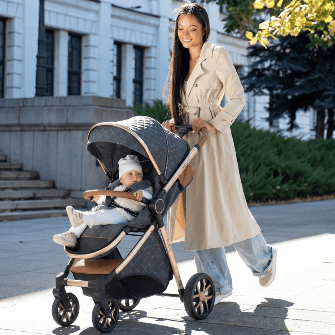 Poussette bébé confortable et design avec cadre doré, canopy matelassé gris et finitions en cuir marron, poussée par une femme souriante dans un parc de ville.