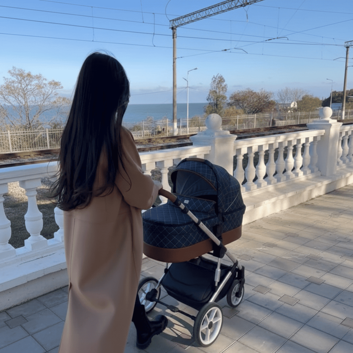 Maman de dos poussant une poussette-landau bébé 3-en-1 chic avec nacelle gris matelassé et détails cuir sur une promenade côtière avec balustrade blanche.